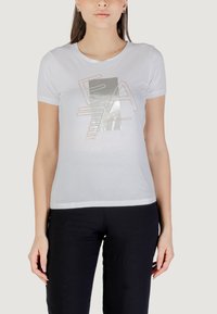 Camiseta ajustada blanca de manga corta. Presenta un texto gráfico metálico en color plata y oro rosa con "EA" y el nombre de la marca "Emporio Armani". Textura de algodón suave.