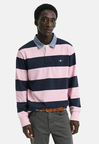 Polo a maniche lunghe a righe blu navy e rosa con colletto in denim, caratterizzato da un piccolo logo sul petto. Indossato con pantaloni grigi e una cintura marrone.
