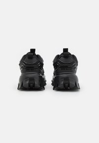 Ein Paar schwarze, klobige Sneakers mit dicken, strukturierten Sohlen und Zuglaschen, von hinten auf einem schlichten weißen Hintergrund betrachtet.
