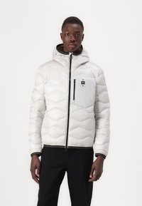 Giubbotto imbottito bianco con cappuccio, dotato di chiusura con zip, tasche frontali e polsini a coste. La texture è trapuntata, con accenti neri e verdi.