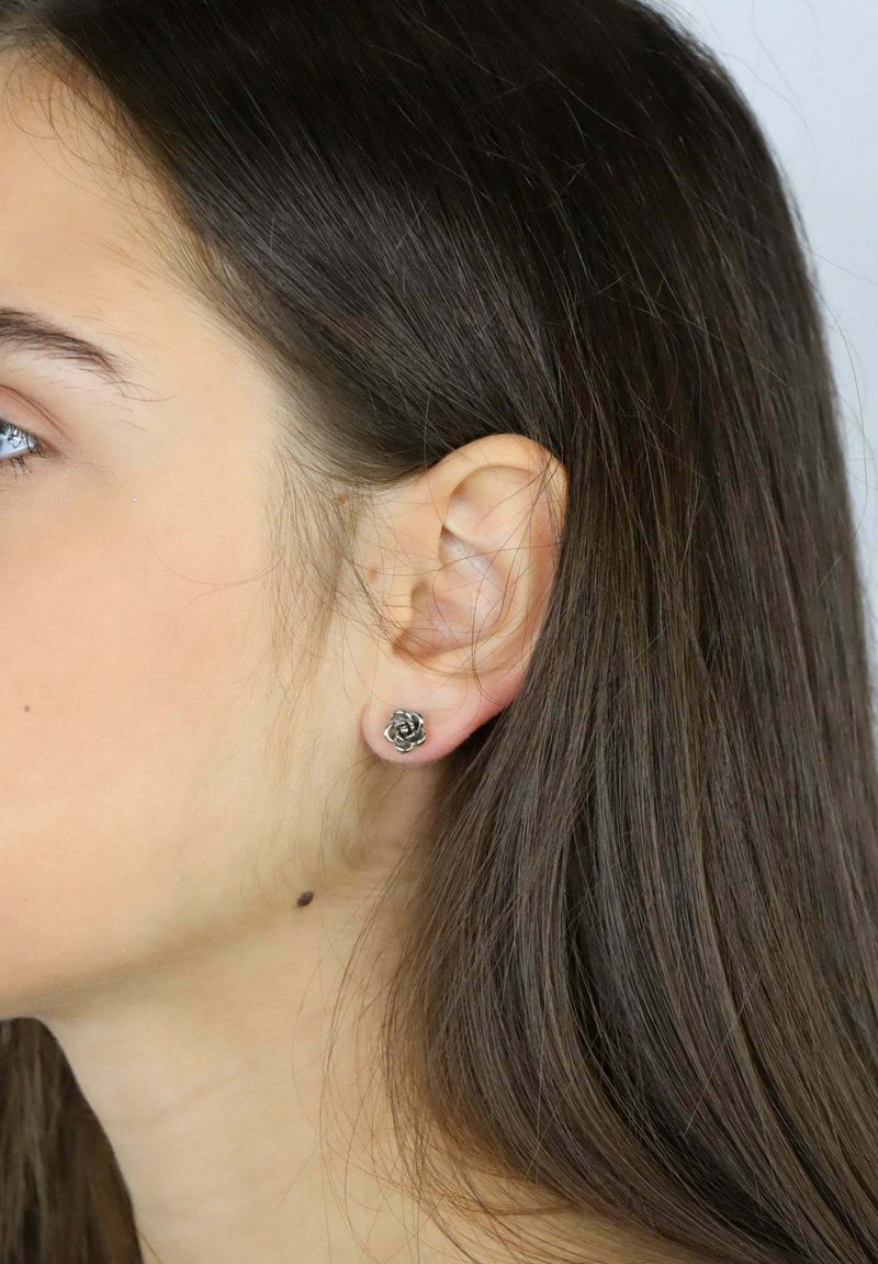 Pendiente de botón en forma de rosa plateada con detalles intrincados, mostrado en la oreja de una modelo junto a un cabello oscuro y liso.