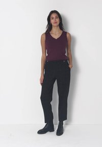 Femme debout contre un mur blanc, portant un haut sans manches violet foncé, un pantalon noir et des bottines noires avec une main dans la poche.