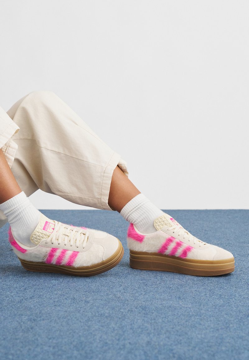 Joggesko med kremfarget, lodden overflate, rosa striper og beige platåsåler. Har en strukturert overdel og polstret tunge, brukt med hvite sokker.