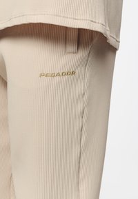 Beige ribbade byxor av tyg med en strukturerad yta, med en broderad "PEGADOR"-logotyp i guld på vänstra låret.