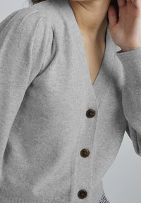 Grå stickad cardigan med V-hals, puffärmar och fem bruna knappar fram. Mjuk textur med en figurnära ribbad fåll.