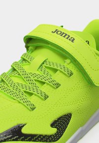 Αθλητικό παπούτσι νέον πράσινο με μαύρα κορδόνια με σχέδιο, μαύρο λογότυπο στη πλάγια πλευρά και ιμάντα με αυτοκόλλητο Velcro με την ένδειξη «Joma» στο πάνω μέρος.