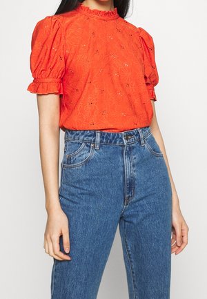 Femme portant un chemisier ajouré à fleurs orange vif avec des manches courtes bouffantes à volants et un jean en denim bleu taille haute.