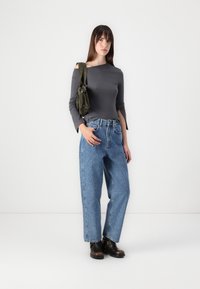 Top gris de manga larga con corte off-shoulder, jeans de talle alto y baggy de color azul, y botas de tobillo marrón oscuro. Modelo sujetando un bolso de mano verde oliva.