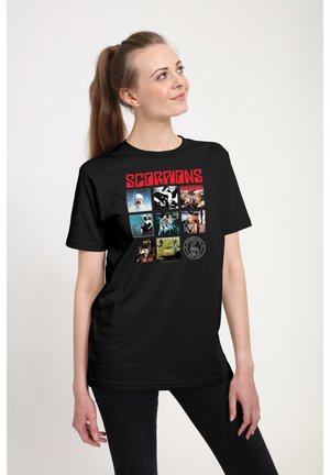 Woman with long brown hair in ponytail wears black Scorpions band t-shirt with album covers, looking slightly up and to the side, against white background.