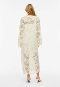 Femme aux longs cheveux portant une robe en dentelle crème transparente à manches longues et des sandales blanches à talons, debout devant un fond blanc.