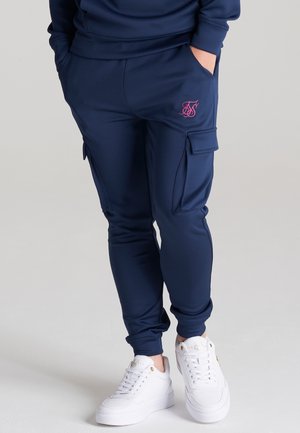 Osoba nosząca granatowe spodnie joggery typu cargo z różowym logo na kieszeni i białe trampki, ręce w kieszeniach, stojąca na jasnej podłodze.