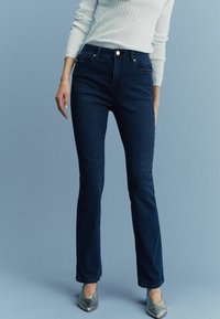 Jeans evazati de culoare bleu închis, cu talie ajustată, cinci buzunare și accesorii din metal auriu. Asortate cu balerini metalici cu vârf ascuțit.