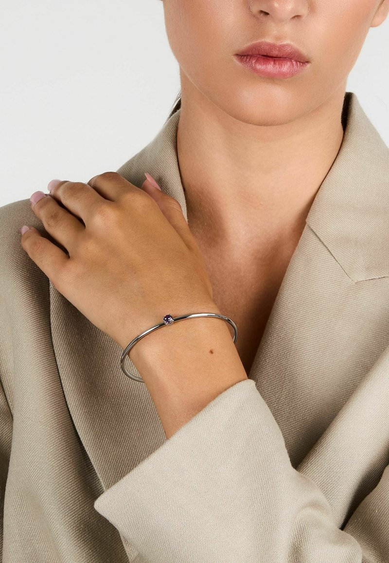 Bracciale in argento con una gemma viola, indossato su una mano appoggiata su un blazer di colore neutro. Finitura in metallo liscia e design minimal.