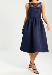 Kvinna i en ärmlös marinblå midiklänning med spets- och paljettobero, hållande en matchande clutch och bärande klackskor med remmar.