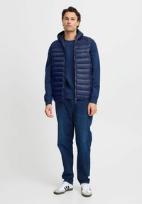 Tumš zilā pūkains bezmugurs veste ar kapuci, quiltēta dizaina, ar priekšējo rāvējslēdzēju, valkāta virs atbilstoša tumš zilā kapuces krekla un zilu džinsu, papildināta ar baltām sporta kurpēm.