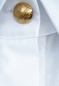 Biała bawełniana tkanina z teksturowanym wykończeniem, ozdobiona złotym, tłoczonym guzikiem z dekoracyjnymi detalami przy dekolcie.