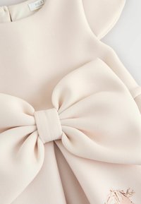 Nærbillede af et blegebeige tøjstykke med en stor stofsløjfe og en diskret trykt illustration nær kanten.