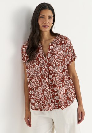 Frau mit langen dunklen Haaren, die eine rostfarbene Kurzarmbluse mit weißen floralen Paisley-Mustern und weiße Hose trägt und vor einer weißen Wand steht.