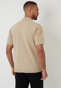 Polo de punto beige de manga corta con un diseño texturizado en las mangas, dobladillo acanalado y cuello. Combinado con pantalones negros.