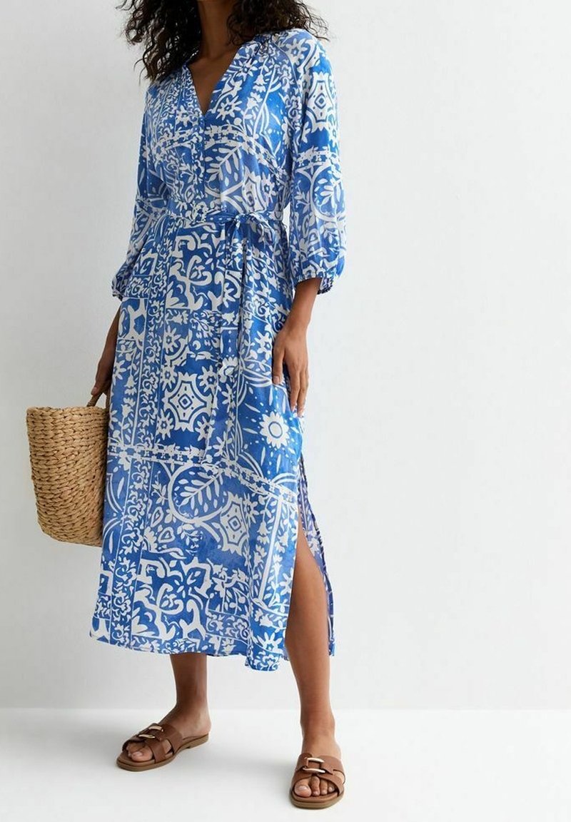 Femme portant une robe midi bleue et blanche à motifs avec une fente latérale, tenant un sac en paille tissé, et des sandales plates marron.