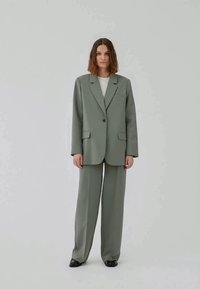 Giacca di suit verde chiaro su misura con revers a scasso, chiusura a un bottone e due tasche frontali, abbinata a pantaloni a gamba larga coordinati.