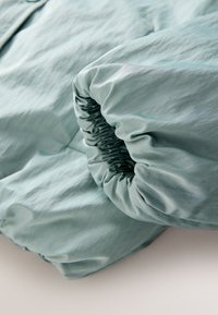 Ljusrått lakanshörn med elastisk kant utsträckt runt madrasshörnet på en slät yta.