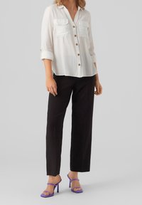 Camicia bianca con bottoni e due tasche frontali; abbinata a pantaloni neri larghi e sandali con tacco viola, creando un look casual.