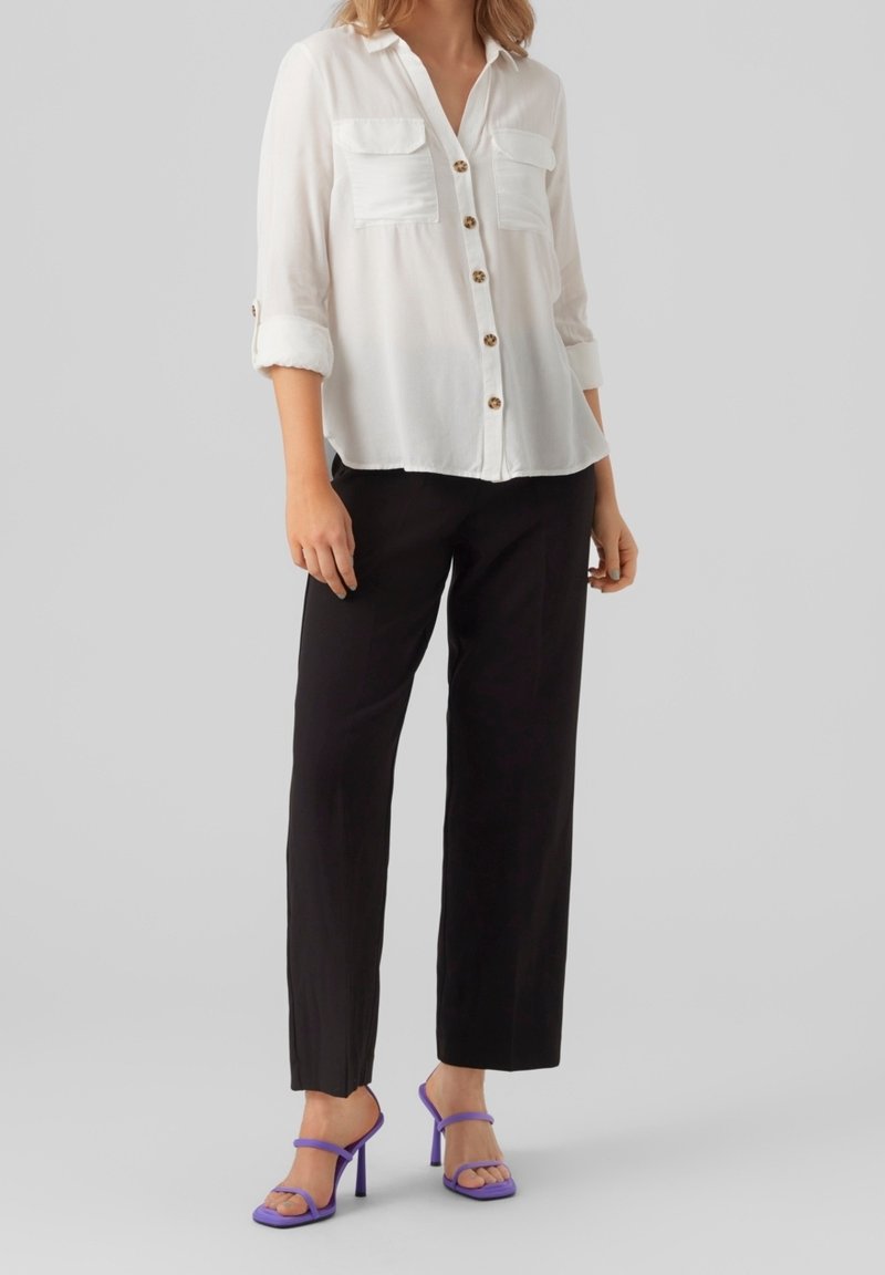 Camicia bianca con bottoni e due tasche frontali; abbinata a pantaloni neri larghi e sandali con tacco viola, creando un look casual.