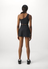 Svart cropped topp med en skulderstropp kombineres med et svart skjørt. Modellen har på seg sorte athletic sko med hvite sokker. Glatt stoff tekstur.
