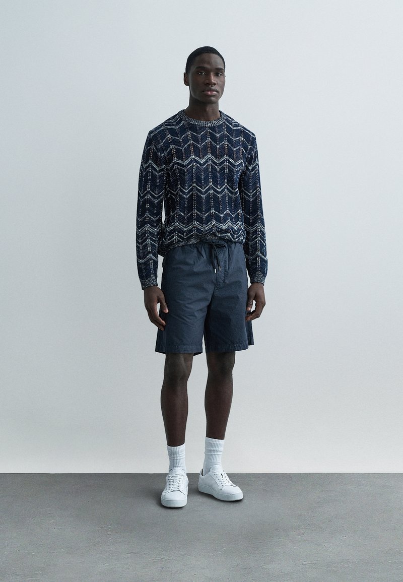 Pull en maille bleu marine à motif zigzag, associé à un short gris, des baskets blanches et des chaussettes blanches montantes. Fond plat et minimaliste.