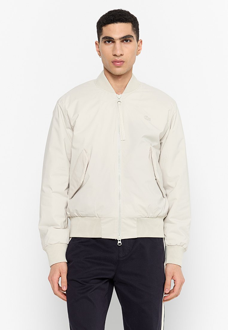 Giacca bomber beige chiaro con zip frontale, polsini e orlo a costine; presenta due tasche laterali e un logo discreto sul petto.