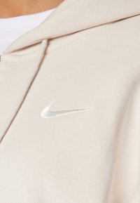 Ljus beige hoodie gjord av mjukt tyg, med en dragsko i huvan. En liten vit Nike-logotyp är broderad på vänster bröst.