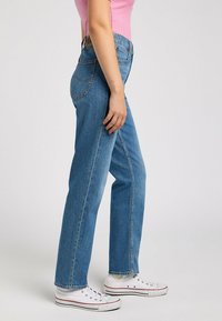 Pantalones vaqueros de denim azul de tiro alto con un diseño de pierna recta, que presentan bolsillos estándar y detalles de costura, combinados con zapatillas blancas.