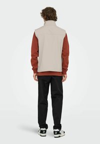 Beige ärmellose Weste über einem roten Langarmhemd, schwarze Hose und schwarz-weiße Turnschuhe. Glattes Material, hoher Kragen und seitliche Taschen.