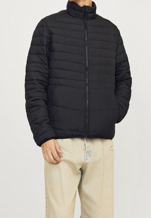 Homme portant une veste noire matelassée zippée avec col montant et un jean beige clair usé avec quelques déchirures.