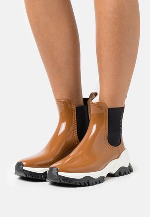 Stivaletti alla caviglia marroni lucidi in gomma, caratterizzati da un pannello laterale elastico nero e una suola spessa in gomma bianca e nera con texture.