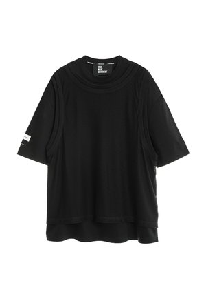 Camiseta negra de manga corta con una superposición sin mangas de malla negra suelta y un pequeño logo blanco en la manga izquierda.