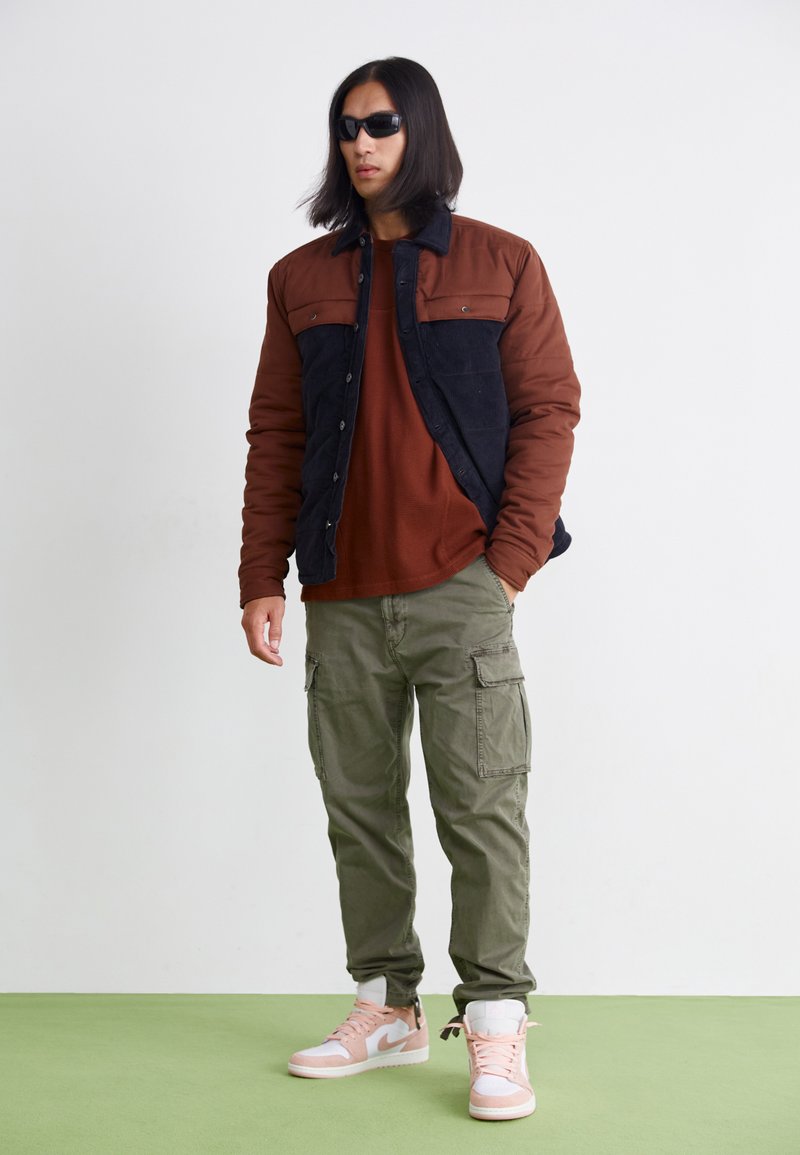 Veste matelassée marron et bleu marine, pantalon cargo olive et baskets roses/blanches. Le mannequin porte des lunettes de soleil noires et se tient devant un fond clair.