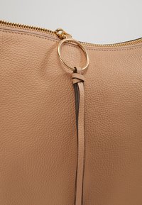 Tan leather handbag featuring a textured finish, gold zipper, and a decorative ring with a braided leather accent.