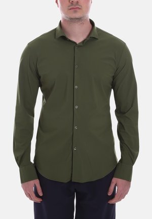 Uomo che indossa una camicia verde oliva aderente a maniche lunghe con bottoni e pantaloni scuri, in piedi contro uno sfondo bianco uniforme.