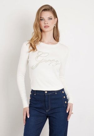 Maglia a maniche lunghe color crema con logo "Guess" in strass, abbinata a jeans in denim scuro a vita alta con dettagli in bottone dorato.