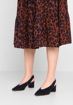 Jambes portant une jupe à imprimé léopard marron et noir et des chaussures à talons blocs slingback en daim noir sur un sol gris.