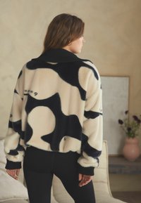 Kreemivärvi valehalli lühike sviiter mustade abstraktsete mustritega. Omab musta soonikraega kaelust ja varrukaotste auku, lõtv sobivus ja pehme tekstuur.