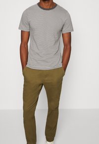 Man in een zwart-wit gestreepte T-shirt met korte mouwen en olijfgroene chino's, met zijn handen in zijn zakken, staand tegen een effen achtergrond.