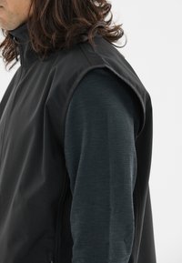 Schwarze ärmellose Jacke aus glattem Stoff, mit hohem Kragen und seitlichen Reißverschlusstaschen, getragen über einem strukturierten, dunkelgrauen Langarmshirt.