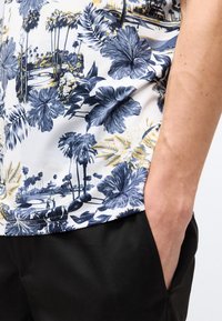 Πουκάμισο με λουλουδάτο και φοινικόδενδρο μοτίβο σε λευκό, μπλε και κίτρινο, φορεμένο με μαύρο παντελόνι, με το χέρι άνετα τοποθετημένο στην τσέπη.