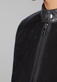 Svart mockajacka med hög krage, dragkedja och tryckknappar vid halsen. Slät textur med vinklade skärningar.