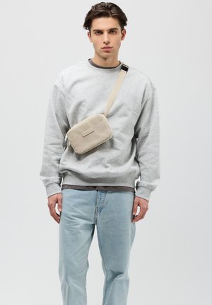 Sac à bandoulière beige en matériau texturé, avec une poche plate à l'avant et une bandoulière ajustable. Porté sur un sweat-shirt gris et un jean bleu.