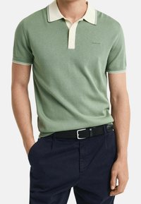 Polo vert avec col blanc et rayures contrastantes, manches courtes, en tissu tricoté texturé, assorti à un pantalon foncé et une ceinture noire.
