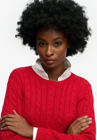 Vrouw met natuurlijk krullend haar draagt een rode kabelgebreide trui over een witte blouse met kraag, met gekruiste armen, die zelfverzekerd naar de camera kijkt.